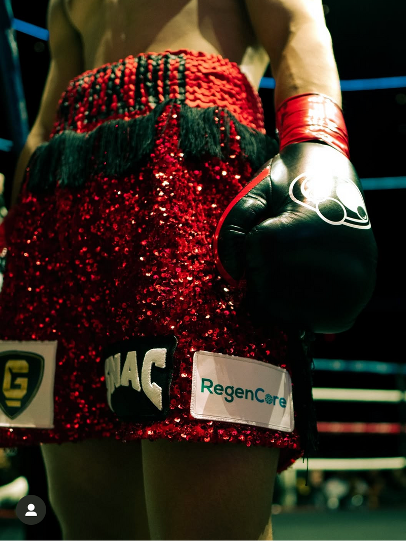 A close-up lifestyle shot of a boxer from behind wearing a red sequined fight skirt with black fringe, red wrist wraps, and black boxing gloves in a boxing ring.